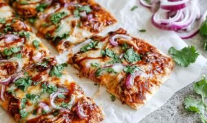 BBQ chicken crust pizza on a baking sheet with melted mozzarella, crumbled bacon, and red onion