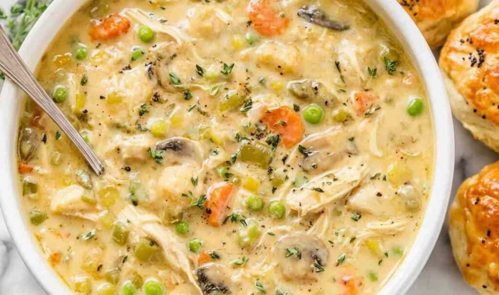 Creamy chicken pot pie soup served in a white bowl with fresh parsley garnish and warm buttered biscuits on the side, showing tender chicken pieces, carrots, and potatoes in a rich broth.