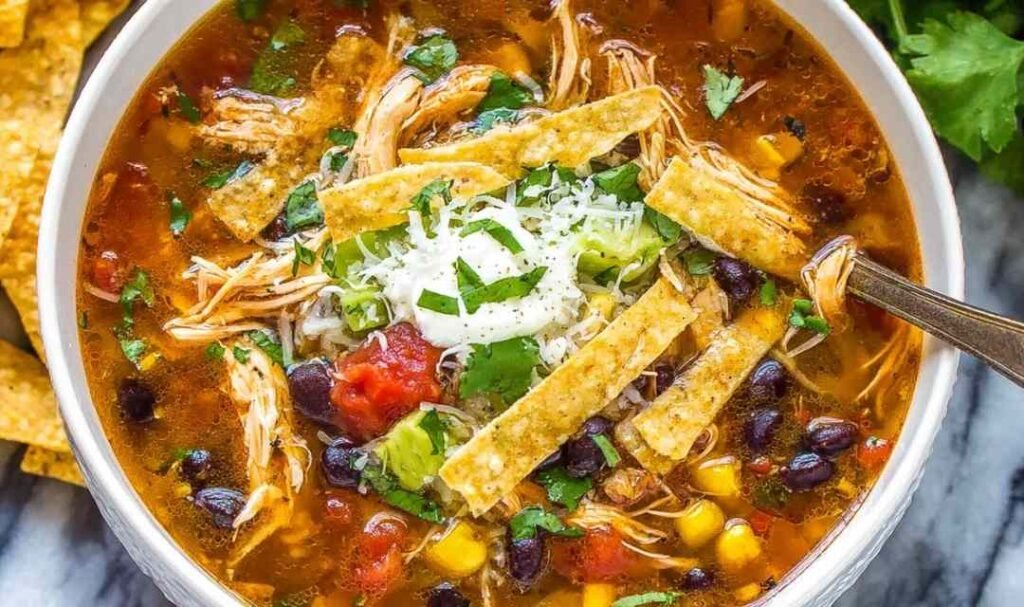 Creamy chicken tortilla soup served in a white bowl topped with crispy tortilla strips, fresh avocado, sour cream, and shredded cheese, showing tender chicken and vegetables in tomato broth.