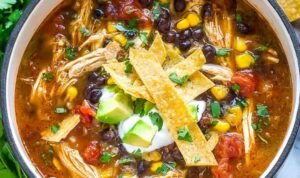 Creamy chicken tortilla soup served in a white bowl topped with crispy tortilla strips, fresh avocado, sour cream, and shredded cheese, showing tender chicken and vegetables in tomato broth.
