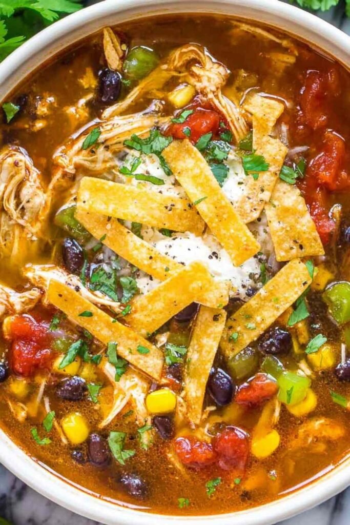 Creamy chicken tortilla soup served in a white bowl topped with crispy tortilla strips, fresh avocado, sour cream, and shredded cheese, showing tender chicken and vegetables in tomato broth.