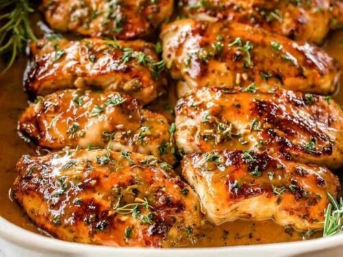 Baked honey mustard chicken breasts in a white baking dish with caramelized glaze and fresh rosemary sprigs