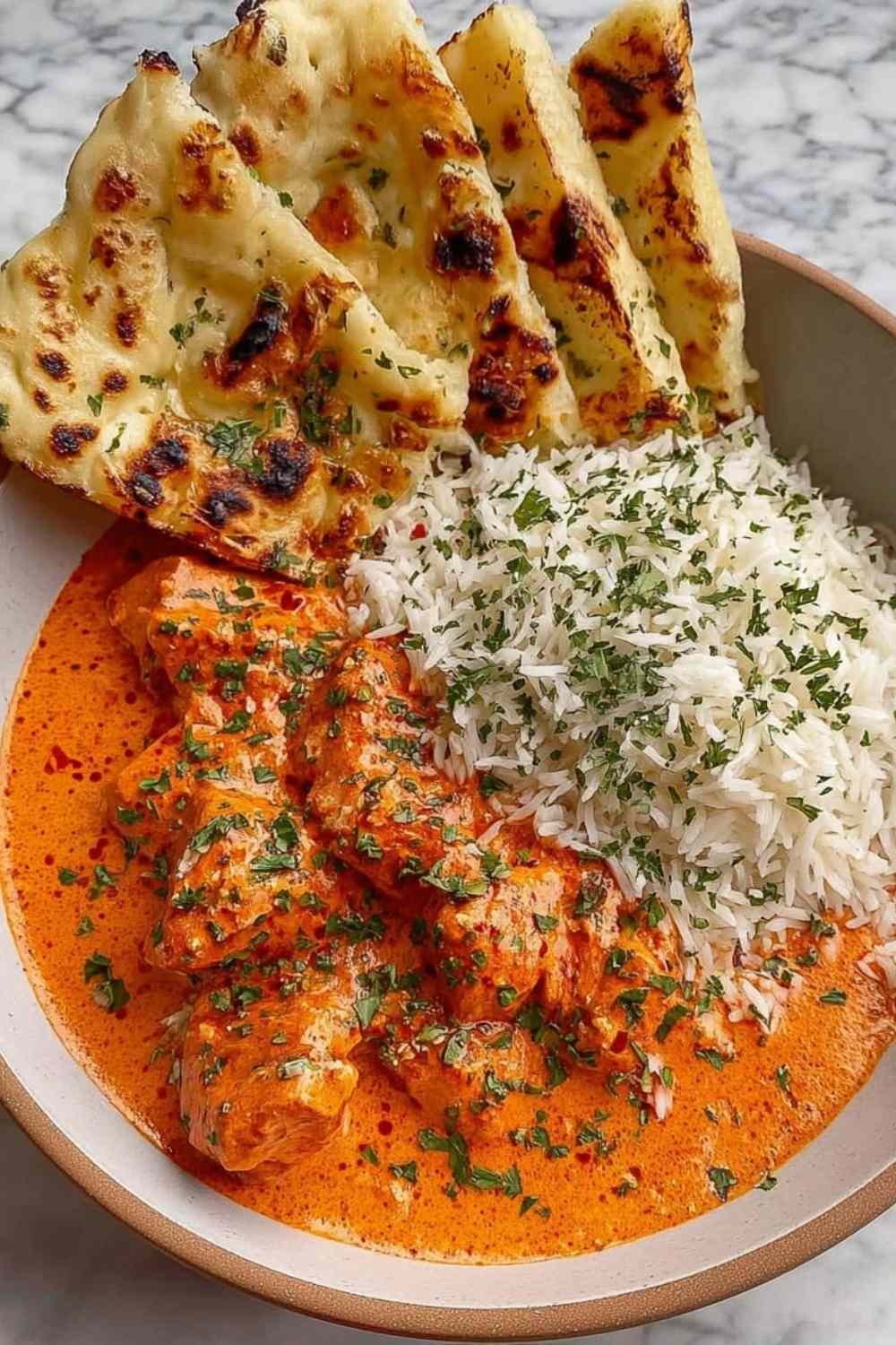 Overhead view of homemade butter chicken recipe in a wide bowl with creamy orange tomato sauce, tender chicken thighs, and fresh parsley garnish alongside warm naan bread
