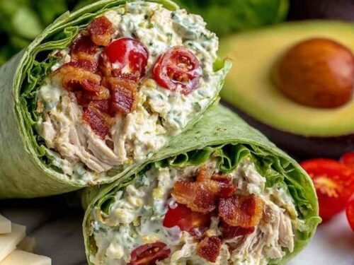 California Club Wraps sliced in half on a wooden cutting board showing layers of crispy bacon, creamy avocado, seasoned rotisserie chicken, and Monterey Jack cheese inside a spinach tortilla