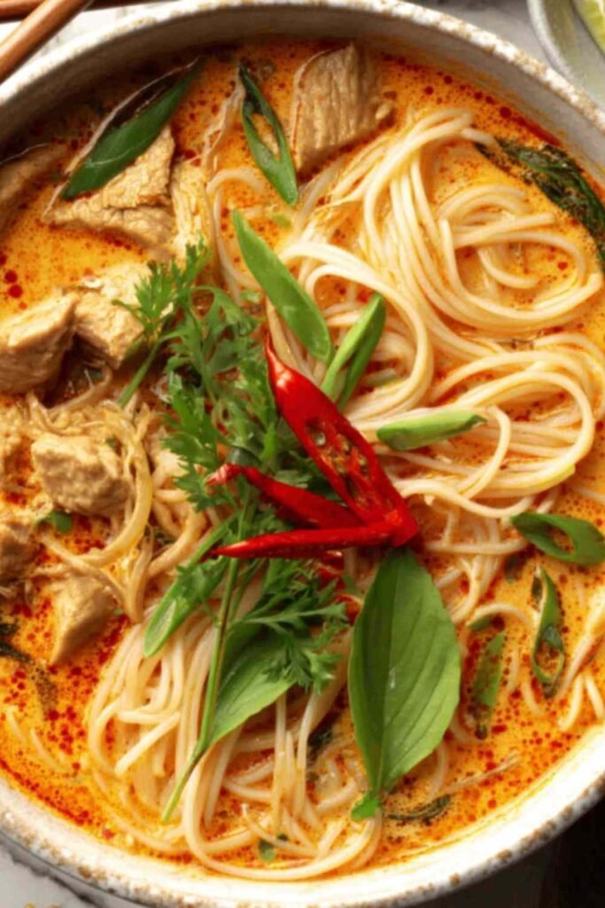 Damn Delicious Thai Red Curry Noodle Soup in a white bowl with fresh cilantro, basil, and a lime wedge garnish