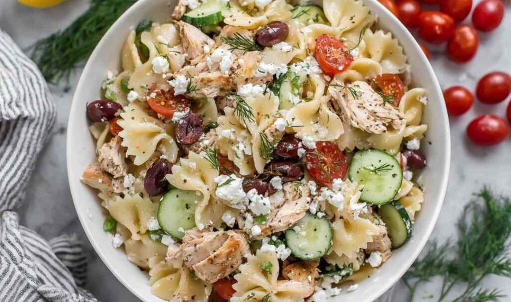 Overhead view of Greek Chicken Pasta Salad with bowtie pasta, cherry tomatoes, cucumber, olives, and feta in a white bowl