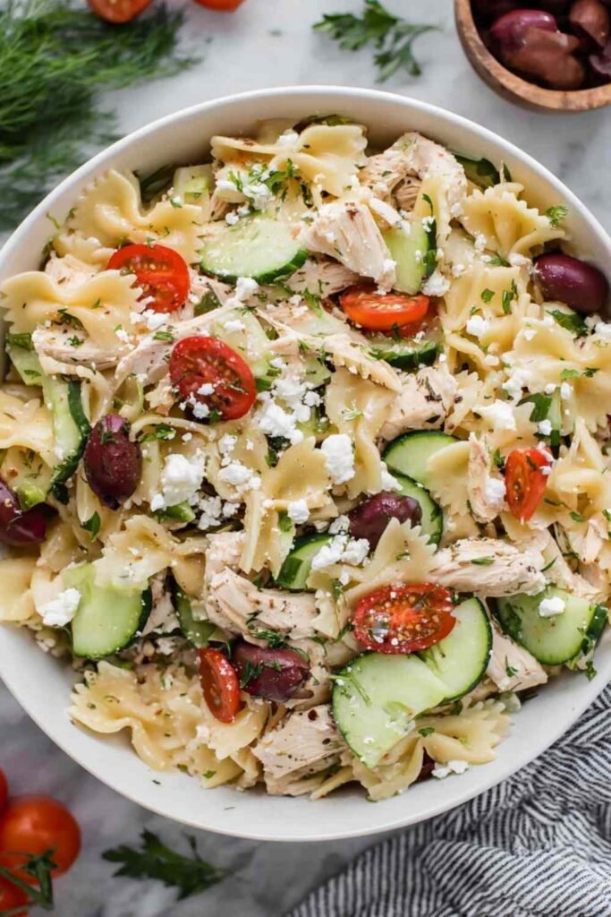Overhead view of Greek Chicken Pasta Salad with bowtie pasta, cherry tomatoes, cucumber, olives, and feta in a white bowl