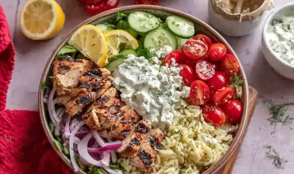 High protein Greek chicken bowls with sliced marinated chicken, rice, fresh vegetables, crumbled feta, and tzatziki sauce in a white bowl