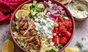 High Protein Greek Chicken Bowls High protein Greek chicken bowls with sliced marinated chicken, rice, fresh vegetables, crumbled feta, and tzatziki sauce in a white bowl