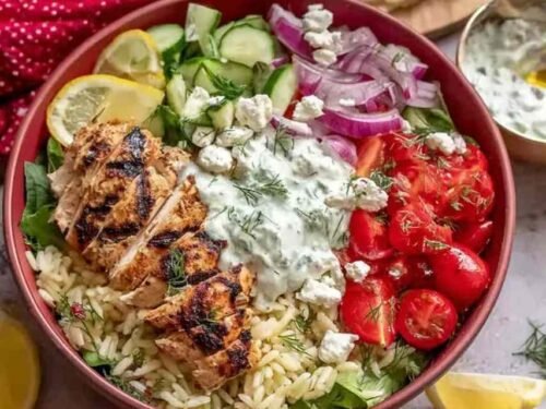 High protein Greek chicken bowls with sliced marinated chicken, rice, fresh vegetables, crumbled feta, and tzatziki sauce in a white bowl