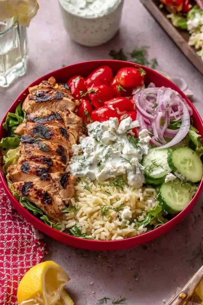 High protein Greek chicken bowls with sliced marinated chicken, rice, fresh vegetables, crumbled feta, and tzatziki sauce in a white bowl