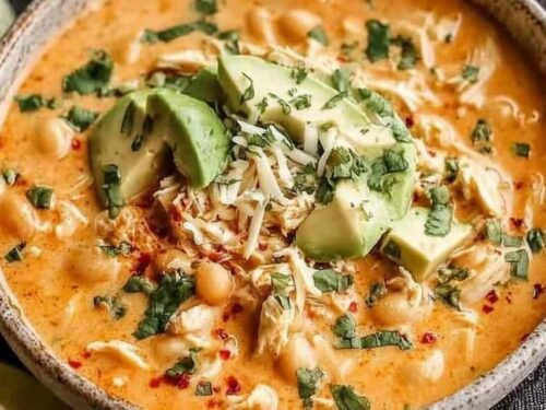 High protein white chicken chili in a white bowl topped with sliced avocado, fresh cilantro, shredded cheese, and a squeeze of lime