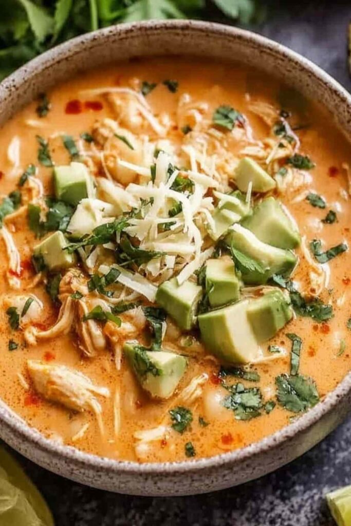 High protein white chicken chili in a white bowl topped with sliced avocado, fresh cilantro, shredded cheese, and a squeeze of lime