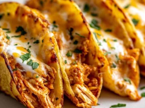Mini baked chicken tacos arranged in a white baking dish with melted cheese and crispy corn tortilla edges