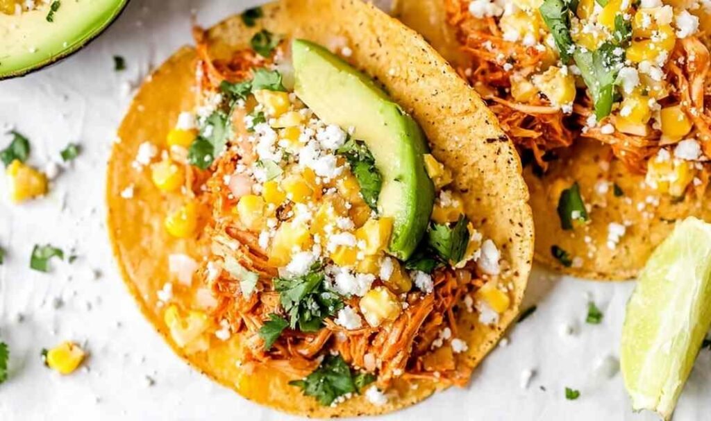 Shredded chipotle chicken tacos topped with corn, avocado, cilantro, and cotija cheese on corn tortillas