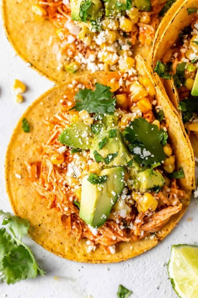 Shredded chipotle chicken tacos topped with corn, avocado, cilantro, and cotija cheese on corn tortillas