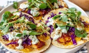 Four smashed chicken tacos on a plate with crispy corn chip crust, melted cheddar cheese, shredded red cabbage, lettuce, and cilantro sauce drizzle