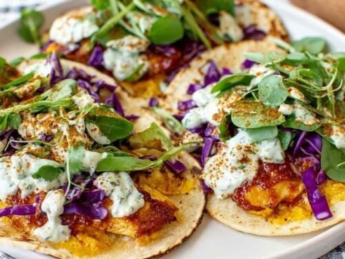 Four smashed chicken tacos on a plate with crispy corn chip crust, melted cheddar cheese, shredded red cabbage, lettuce, and cilantro sauce drizzle